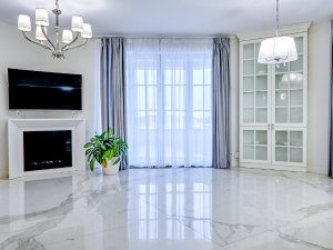 Bright modern living room with a wall-mounted TV above a white fireplace, chandelier, potted plant, and glass-front cabinets beside large windows.